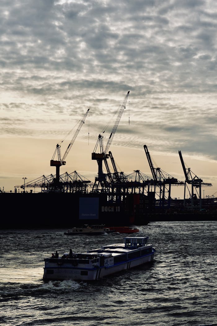 A cargo ship passes a busy industrial port with cranes silhouetted against a sunset sky.